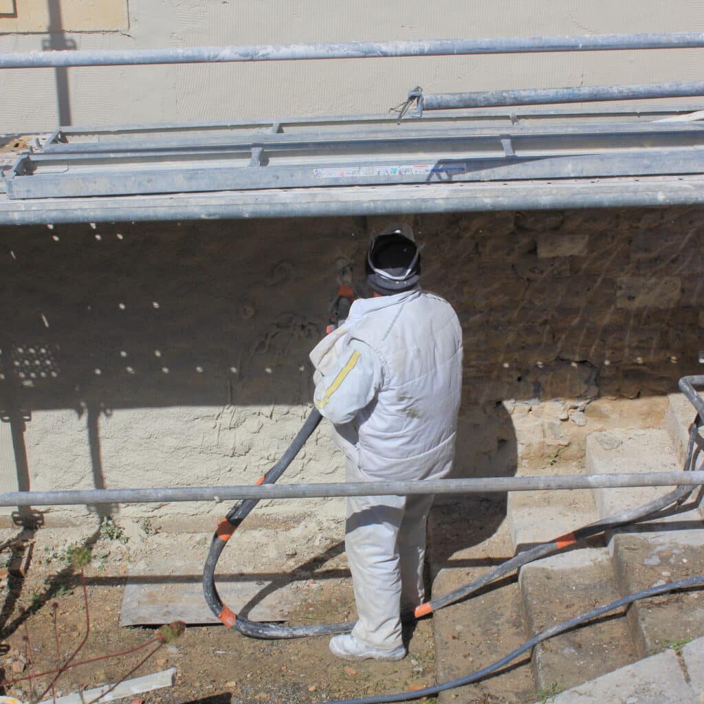 Ouvrier en combinaison blanche appliquant un enduit projeté sur un mur de pierre, sous un échafaudage.