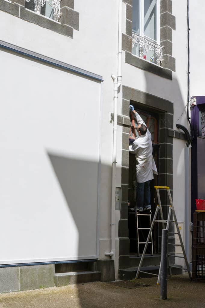Peintre rénovant une façade avec porte sombre Peintre en blanc sur escabeau jaune, ajustant ou peignant l'encadrement d'une porte ou fenêtre d'une façade blanche avec pierres grises.