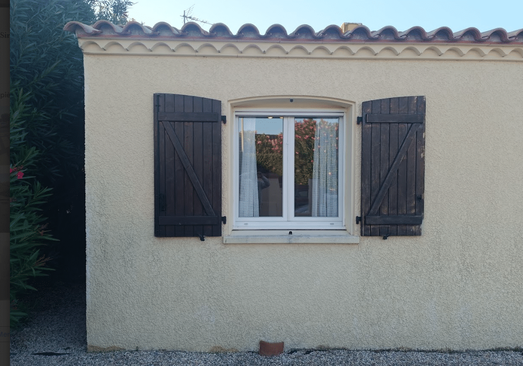 Façade de maison provençale avec fenêtre blanche, volets en bois marron et toit en tuiles romanes sur mur crépi beige.