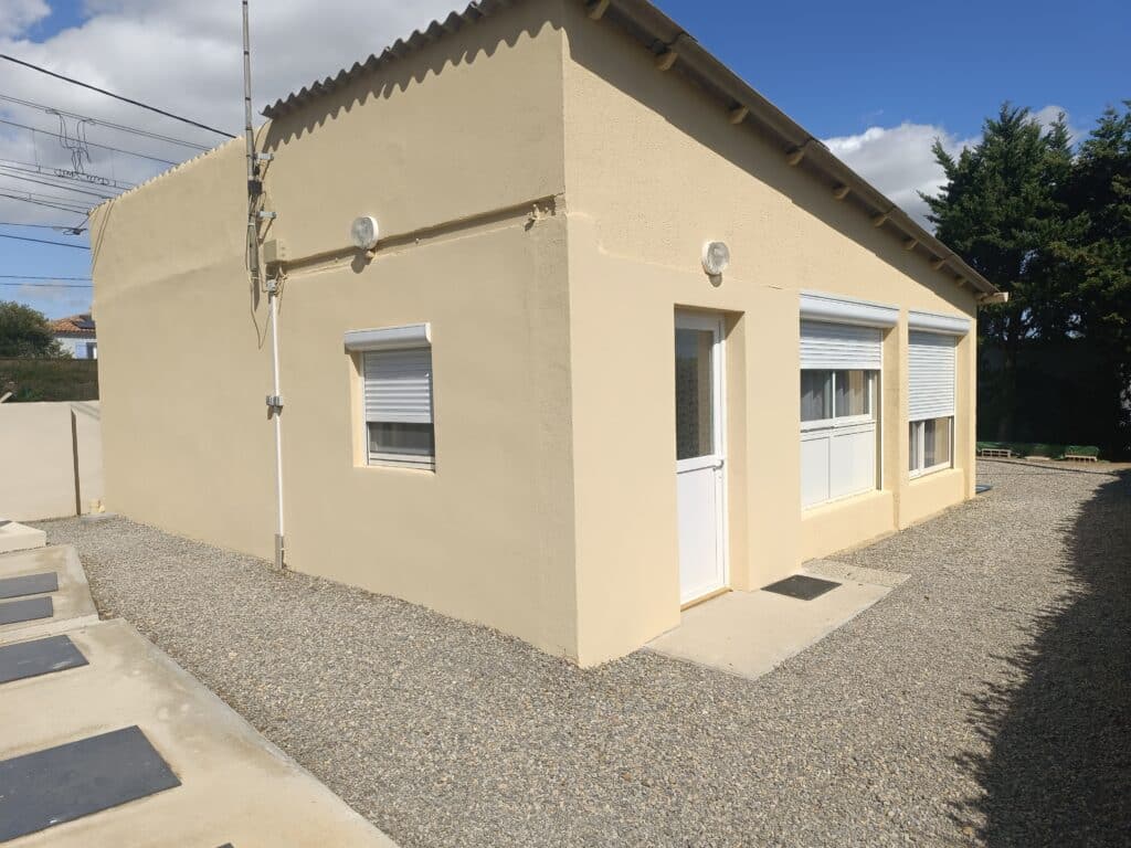 Façade de maison beige avec volets roulants blancs et cour en gravier gris sous un ciel bleu ensoleillé.
