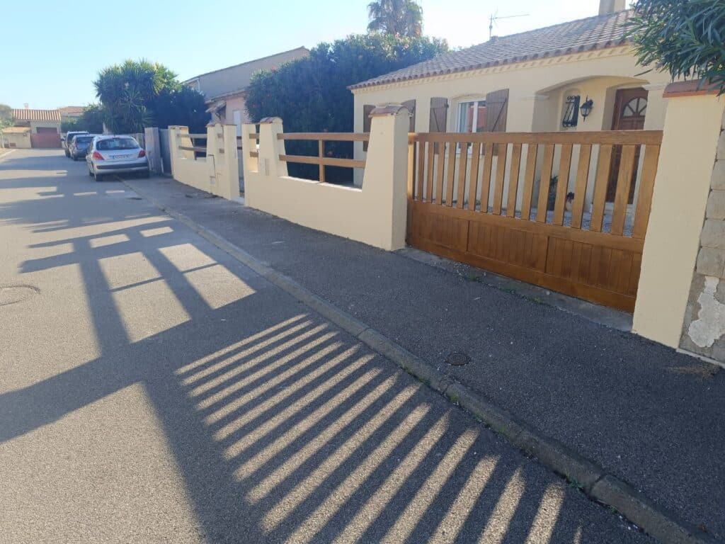 Portail en bois et clôture assortie devant une maison de plain-pied avec ombres projetées sur la rue.