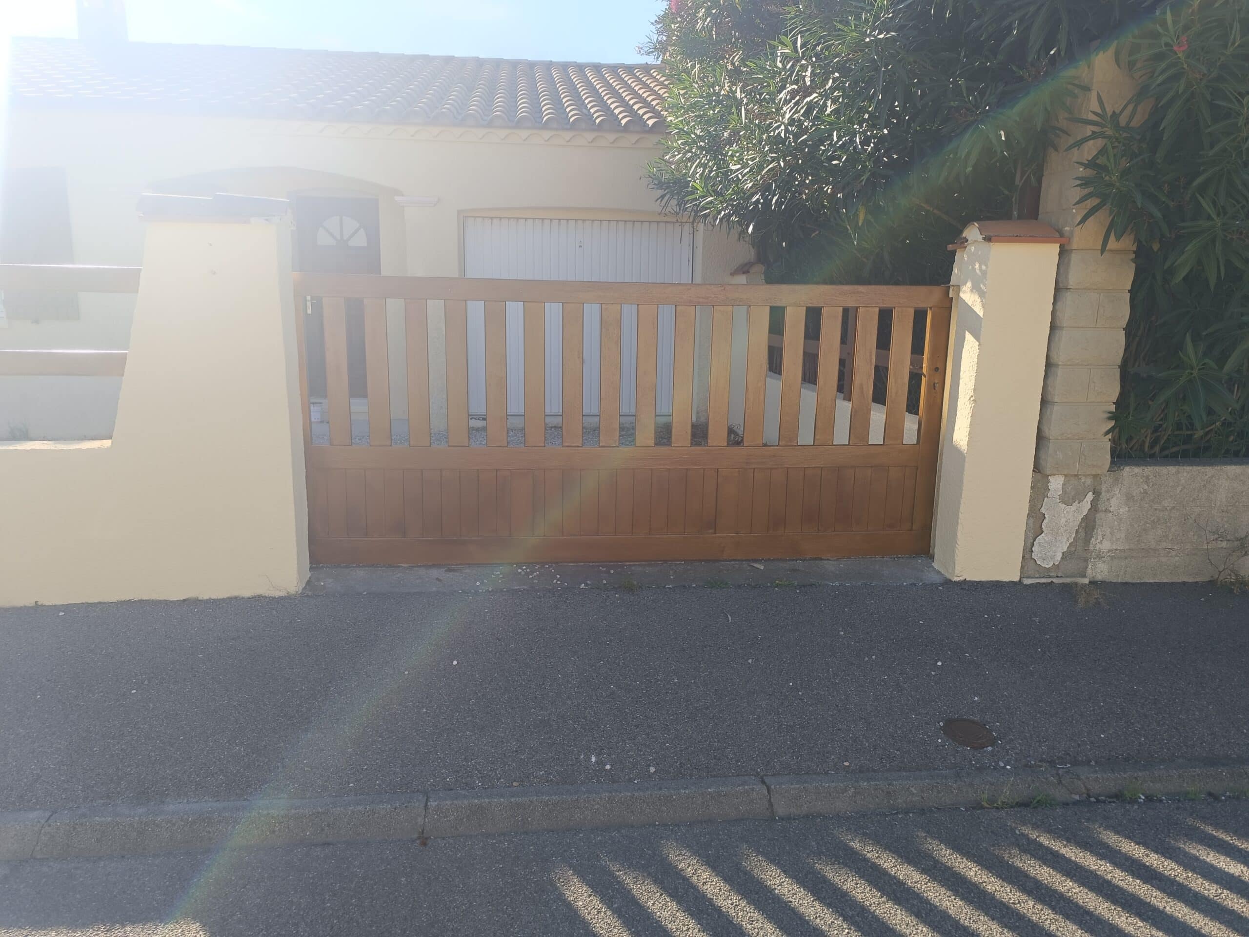 Portail en bois marron devant une maison avec garage blanc, mur de clôture beige et lumière du soleil.