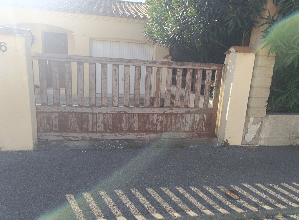 Portail en bois marron à peinture écaillée devant une maison numéro 6 avec garage blanc et arbres.