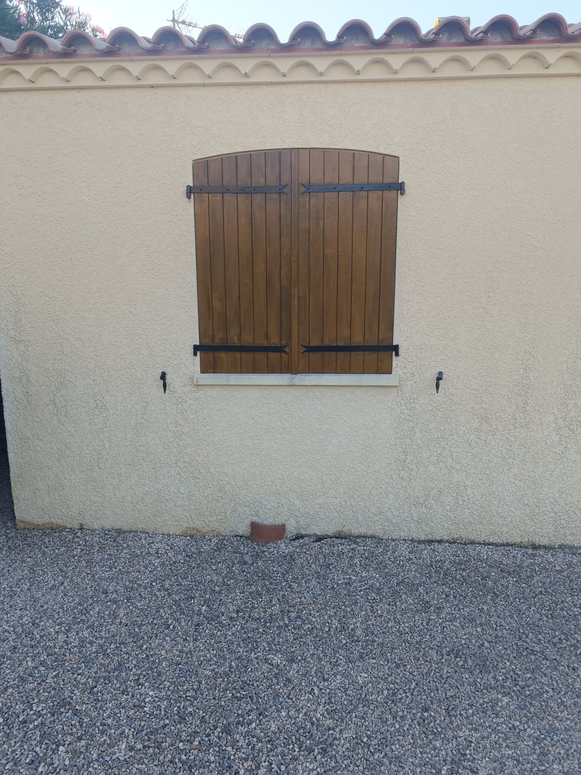 Volet en bois brun fermé sur une façade en crépi beige avec toit en tuiles romanes et sol en gravier.