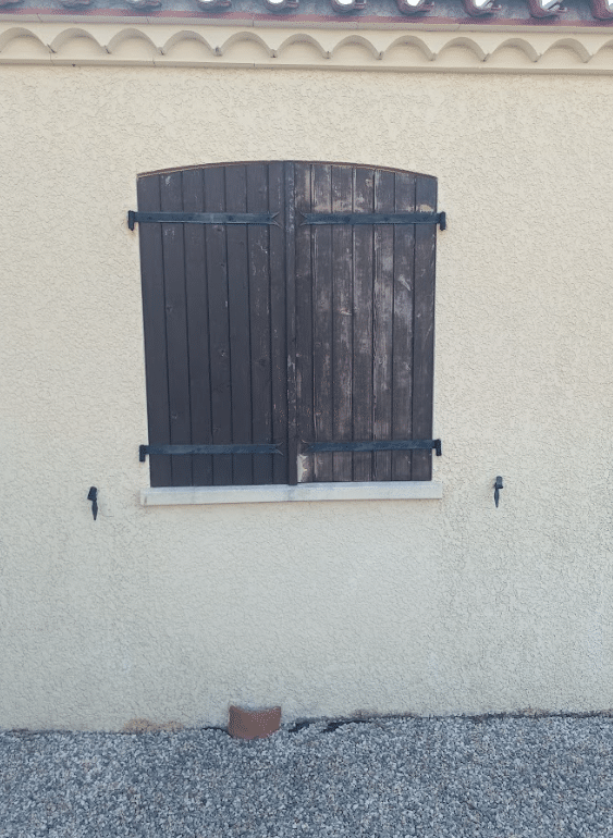 Volets battants en bois marron foncés fermés sur une façade beige avec ferrures décoratives.
